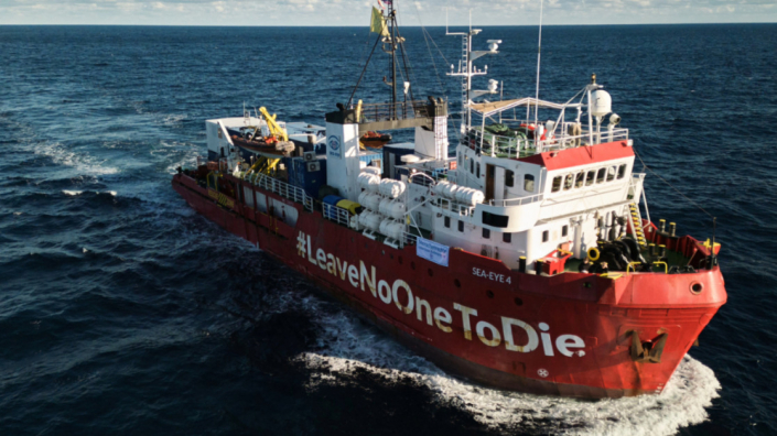 Ein rotes Schiff fährt im Meer. Auf dem Steuerbord steht der Schriftzug "#LeaveNoOneToDie".