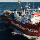Ein rotes Schiff fährt im Meer. Auf dem Steuerbord steht der Schriftzug "#LeaveNoOneToDie".