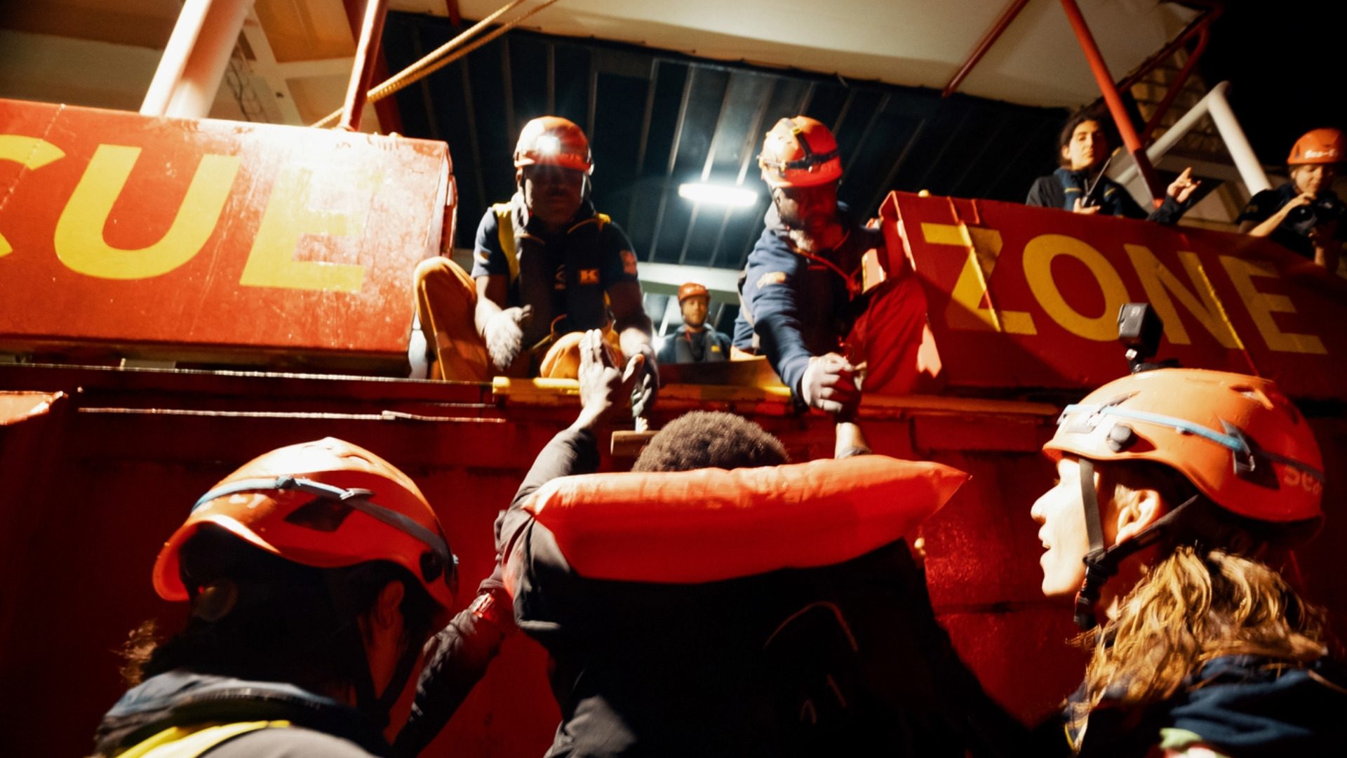 Zwei Menschen stehen am Rand der Rettungszone des Schiffes und geben einer Person mit Schwimmweste die Hand, um sie an Bord zu heben. Zwei Menschen stehen unten und helfen von dort.