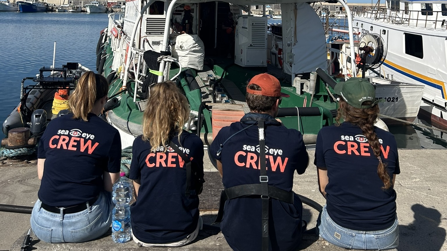 Vier Sea-Eye-Crewmitglieder sitzen mit dem Rücken zur Kamera vor dem Rettungsschiff im Hafen