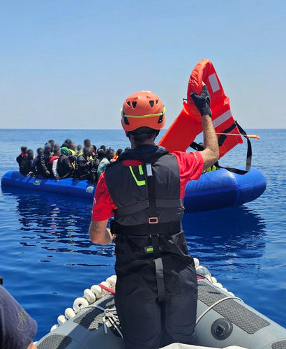 Sea-Eye crew member holds up a life jacket and approaches an overcrowded inflatable boat on the Mediterranean with the RHIB