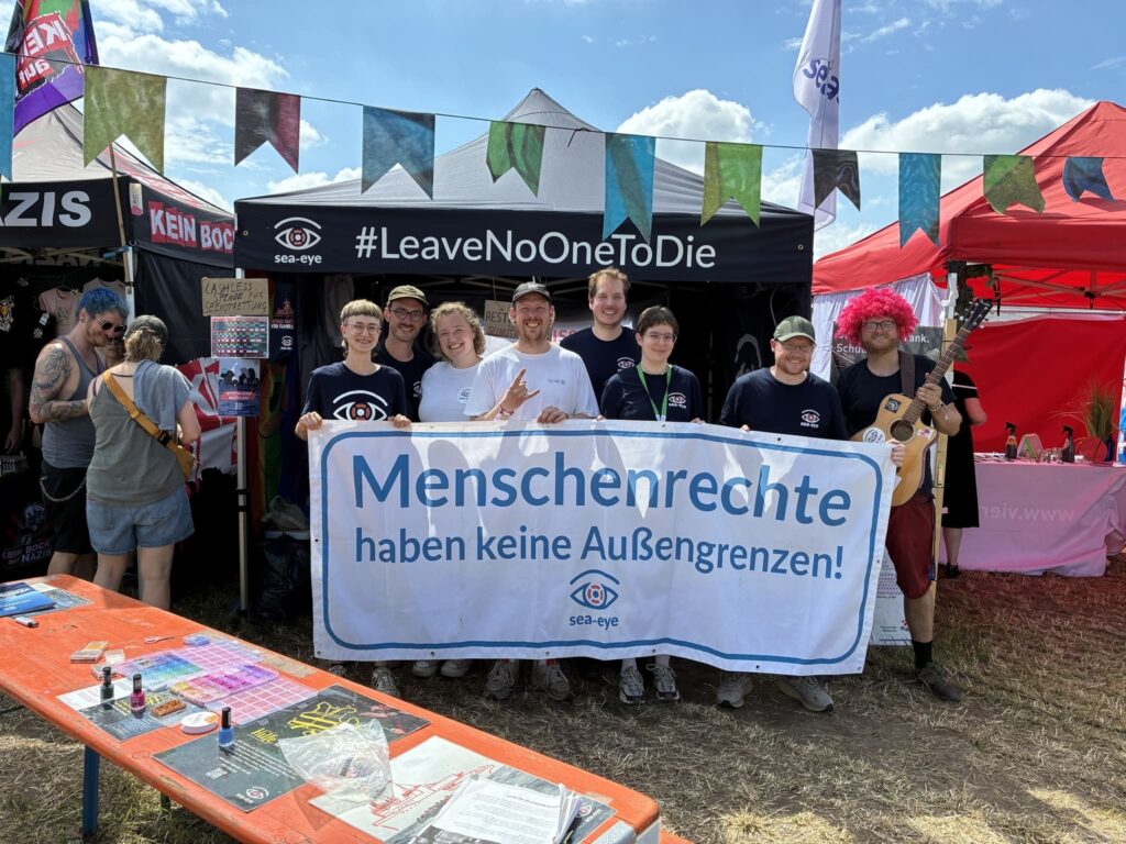 Ehrenamtliche von Sea-Eye zeigen sich vor ihrem Infostand mit einem Banner, auf dem steht: „Menschenrechte haben keine Außengrenzen”.