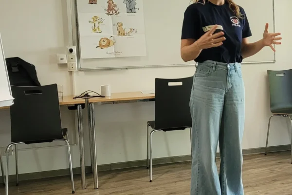 Eine Frau steht vor einem Tisch und mehreren Stühlen in einem Schulungsraum und gestikuliert während eines Vortrags.
