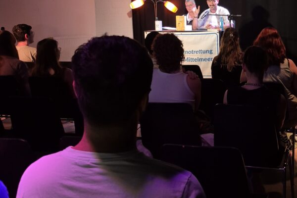 Zwei Personen sitzen an einem Tisch mit Mikrofon und Leselampe vor einem Publikum in einem dunklen Raum