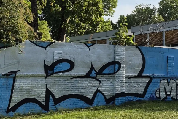 Graffiti mit der Zahl '122 Mio' in großen, schwarz umrandeten Buchstaben auf einer Mauer vor Bäumen und einem Gebäude.