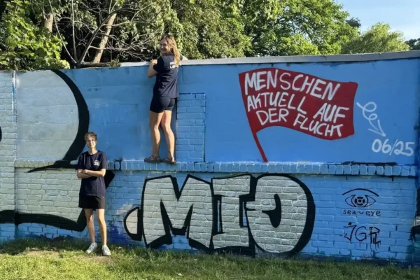 Zwei Ehrenamtliche von Sea-Eye e.V. besprühen eine Wand mit der Zahl '122 Mio' und einem roten Banner mit der Aufschrift 'Menschen aktuell auf der Flucht'