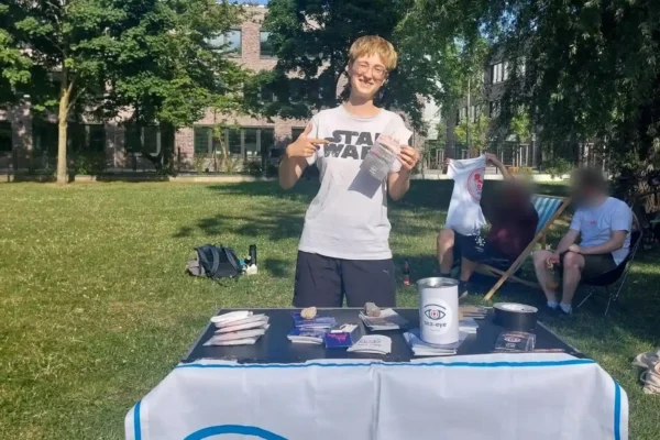 Junge Person steht hinter einem Infostand mit einem Banner von Sea-Eye e.V. und hält Informationsmaterial in der Hand, im Hintergrund weitere Personen auf Stühlen im Park.