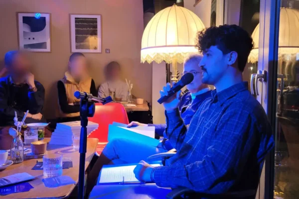 Junger Mann mit Mikrofon und Buch sitzt auf einem Stuhl in einem Raum mit mehreren Zuhörern und einer Stehlampe