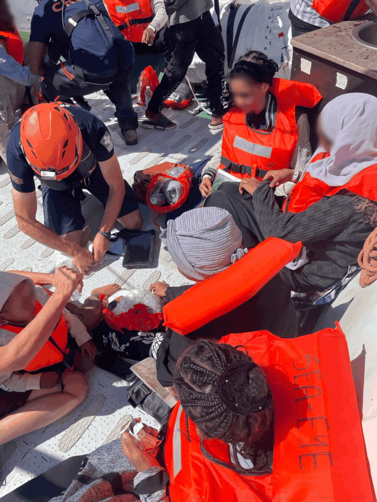 Mehrere Menschen in Rettungswesten sitzen und liegen an Deck eines Rettungsschiffs, während eine Person mit Helm Erste Hilfe leistet