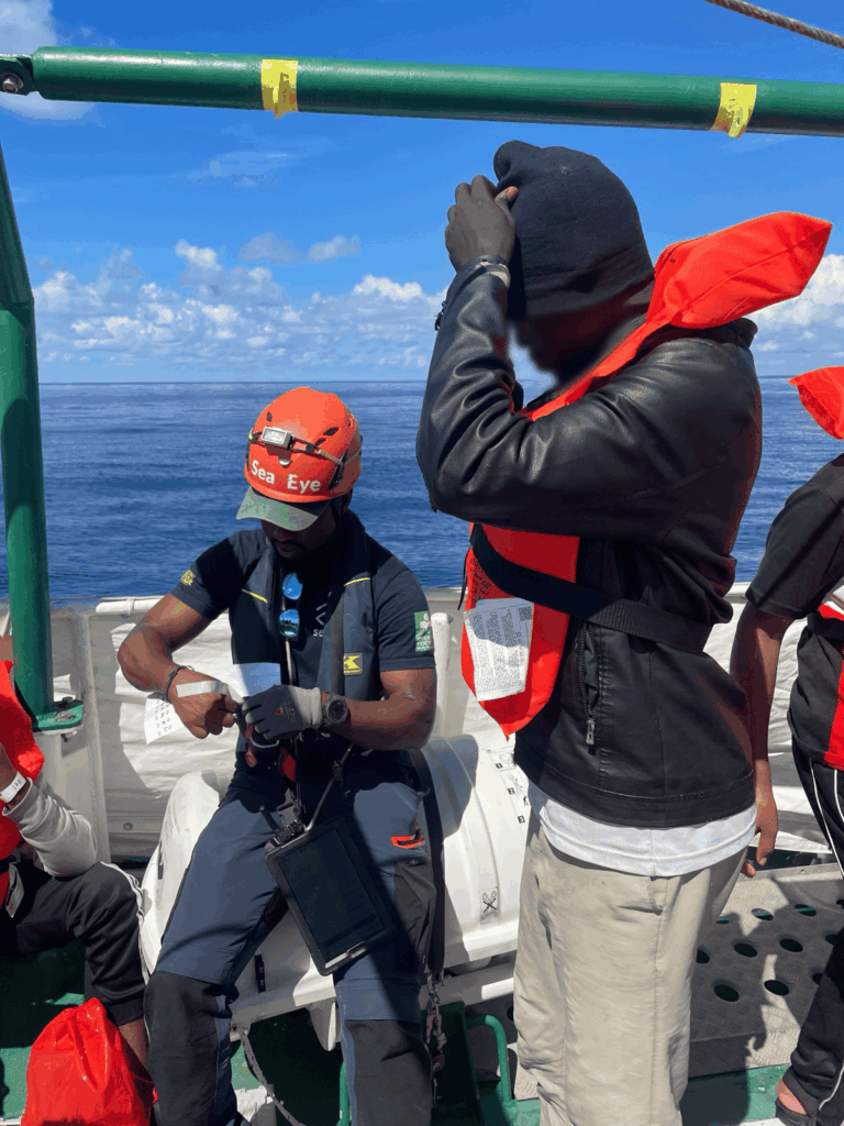 Crewmitglied der SEA-EYE 5 mit oranger Rettungsweste und Helm sitzt auf einem Schiff, daneben steht ein Überlebender mit schwarzer Mütze und oranger Rettungsweste vor dem offenen Meer
