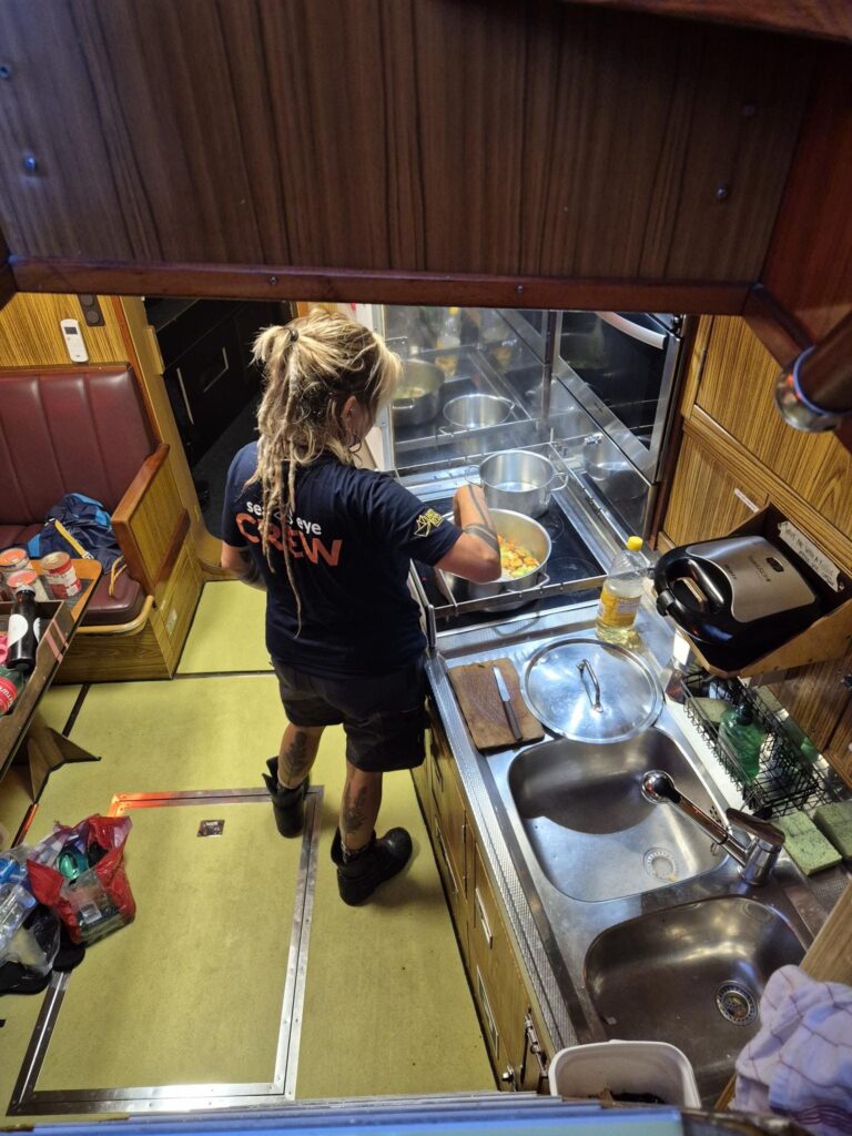 Person mit Sea-Eye-Crew-Shirt steht in einer Schiffsküche und rührt in einem Topf auf dem Herd