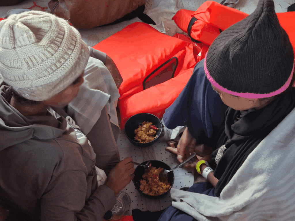 Zwei Menschen sitzen an Bord eines Rettungsschiffes, tragen Mützen und essen aus schwarzen Schalen mit Löffeln.