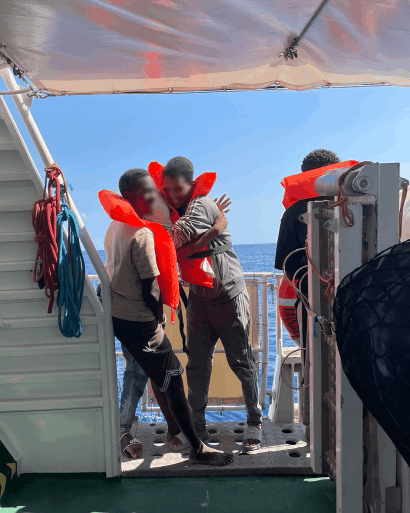 Two rescued people wearing life jackets embrace on a ship deck with open sea in the background.