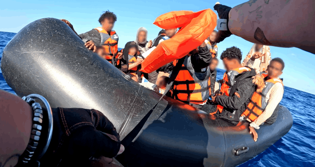 Mitglied des RHIB Teams reicht schutzsuchenden Menschen auf einem Schlauchboot eine orangefarbene Rettungsweste
