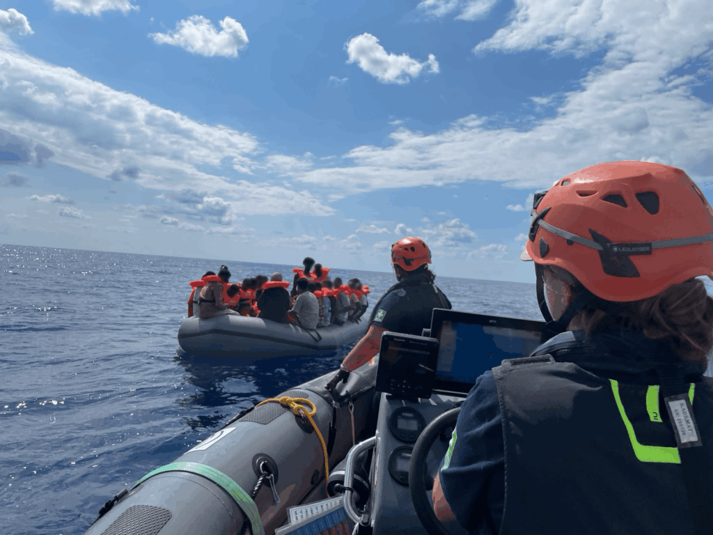 Zwei Boote auf offenem Meer, eines mit Rettungsteam in Schutzhelmen und das andere mit mehreren Menschen in Rettungswesten.